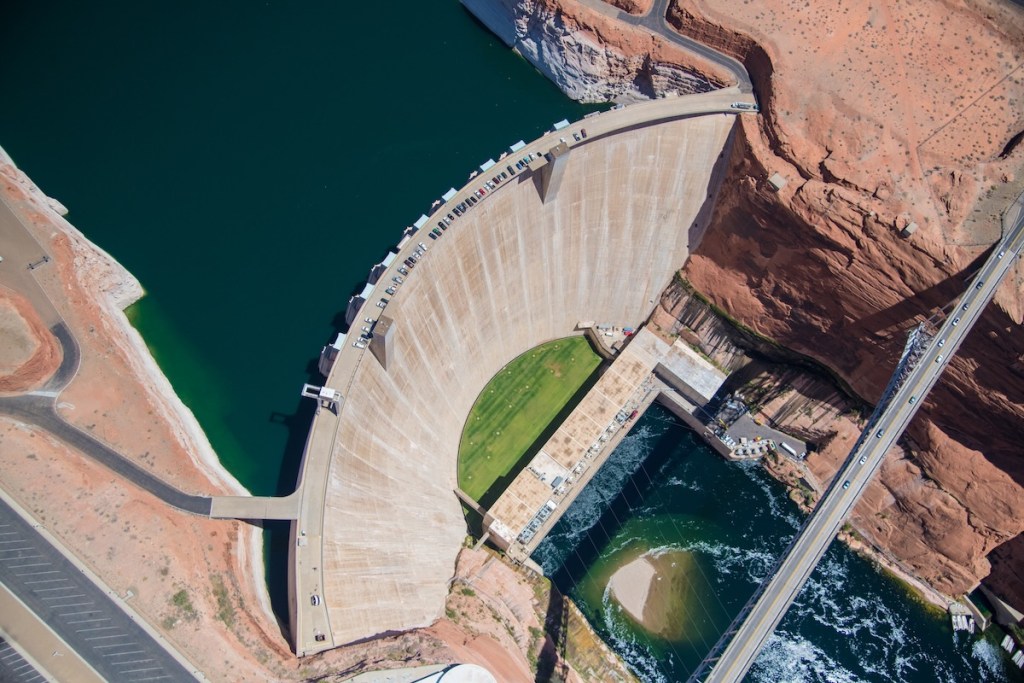 Aerial view of a large concrete dam with a curved structure, surrounded by rocky terrain and a deep blue reservoir on one side.