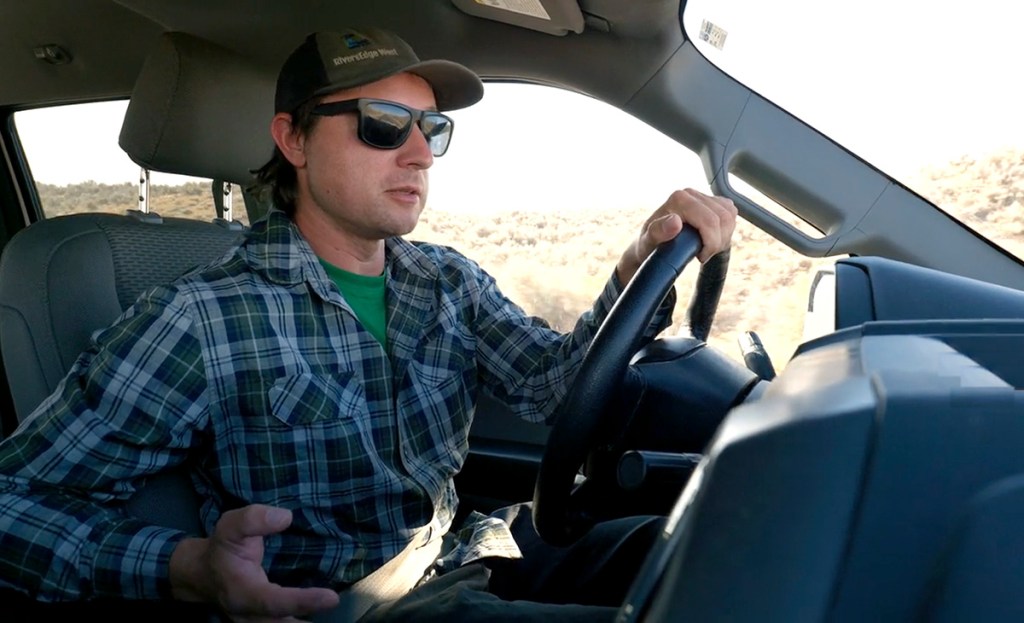 A man driving a vehicle, wearing sunglasses and a plaid shirt, appears focused while steering with one hand.