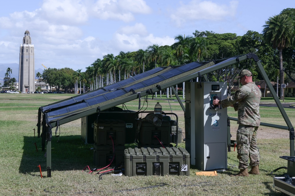 A soldier operating a portable solar power unit with solar panels deployed, near military equipment, in a park-like setting with a tall building in the background.