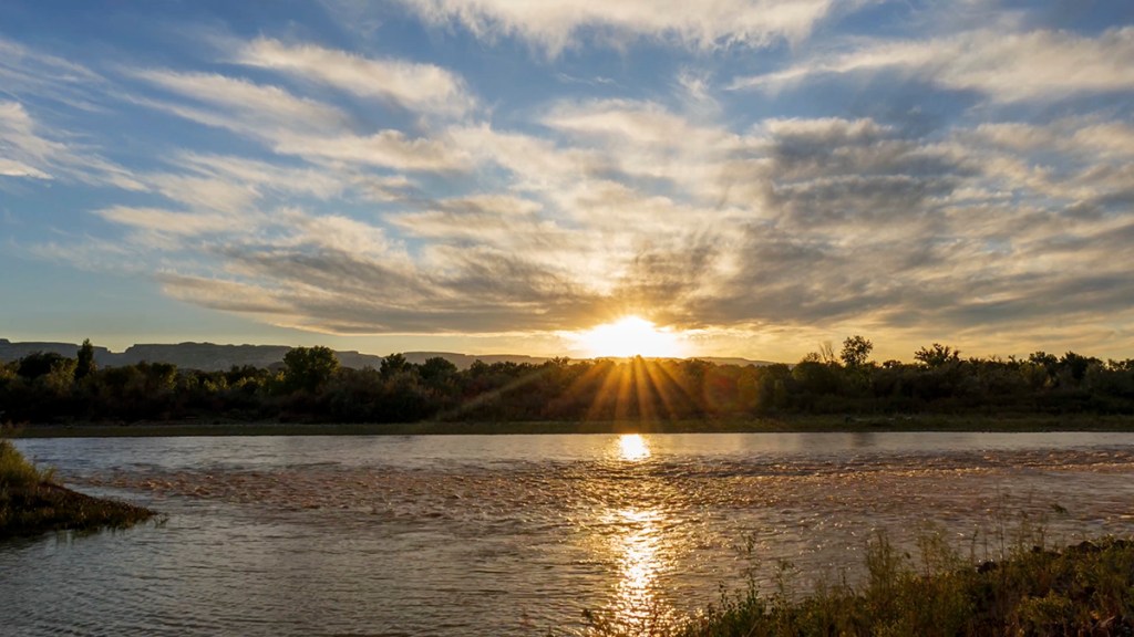 A serene sunset over a river, with golden rays shining through the clouds and reflecting on the water, surrounded by lush greenery.