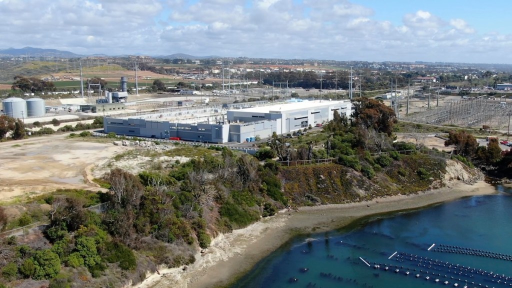 Aerial view of a modern industrial facility located near a body of water, with a surrounding landscape featuring greenery and electrical infrastructure in the background.