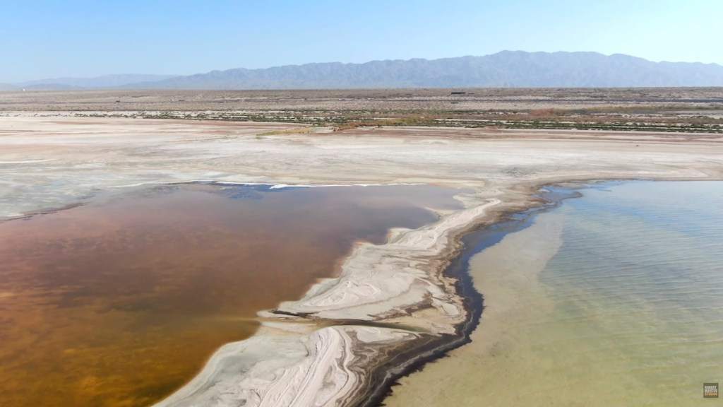 Aerial view of a salt flat with contrasting colors of water and land, surrounded by mountains in the distance.