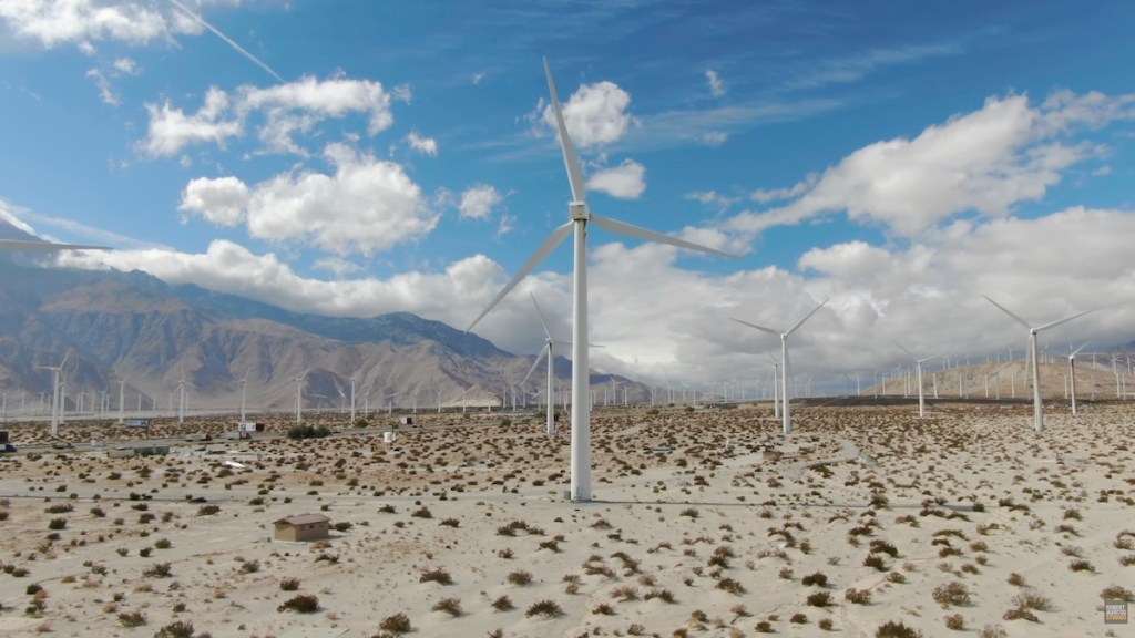 A landscape featuring multiple wind turbines standing in a desert area against a backdrop of mountains and cloudy blue sky.