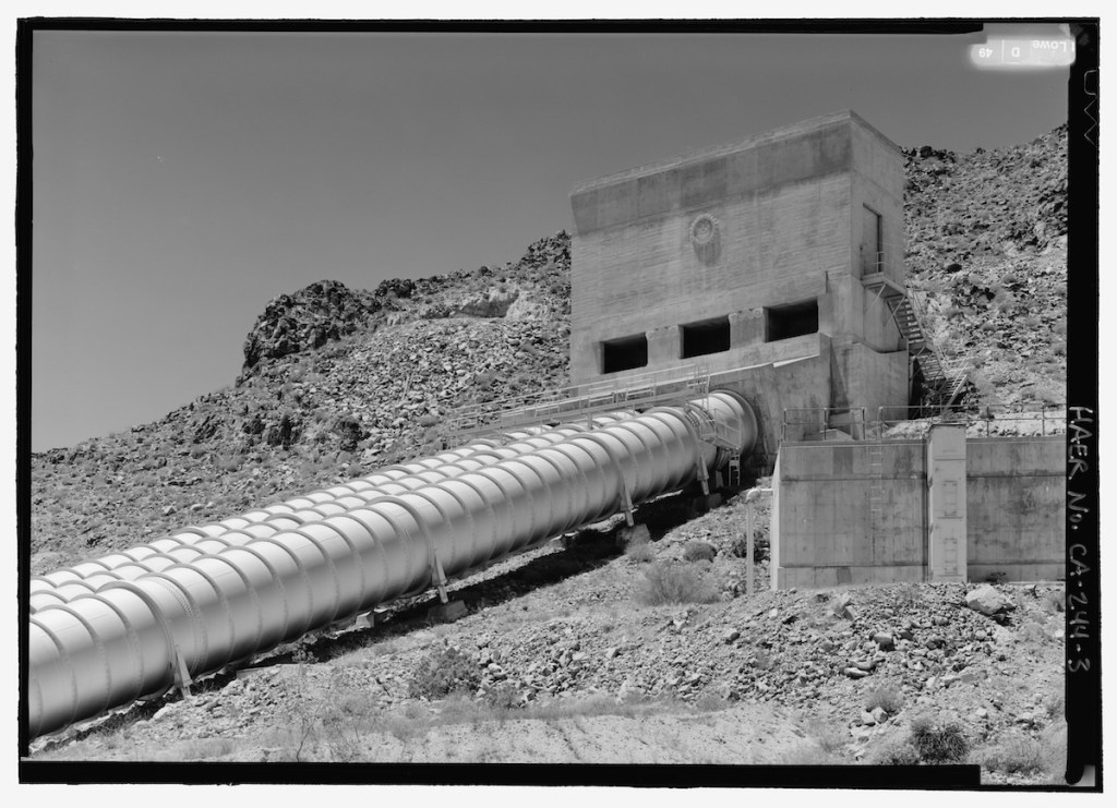 A black and white photograph of a large concrete structure on a rocky hillside, featuring a long, cylindrical pipeline extending from it.