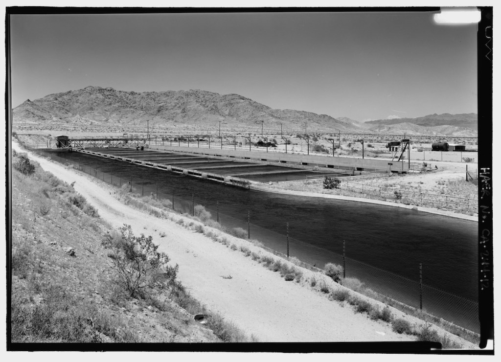 A black and white photograph of a water canal in a desert landscape, with mountains in the background and a fence along the water's edge.