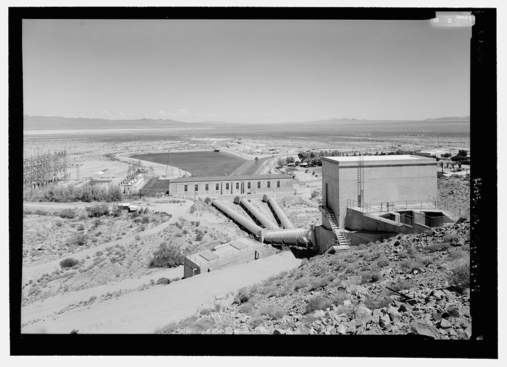 Aerial view of a large industrial facility in a desert landscape, featuring water storage tanks, pipelines, and electrical infrastructure.