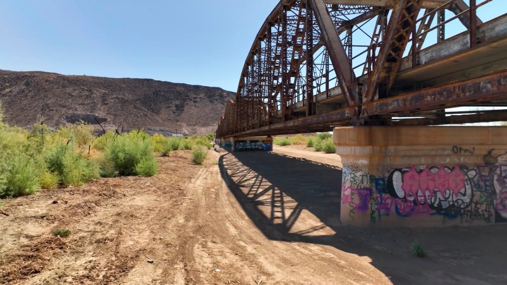 A rusty metal bridge with an arched design, positioned over a sandy path surrounded by sparse vegetation and hills in the background.
