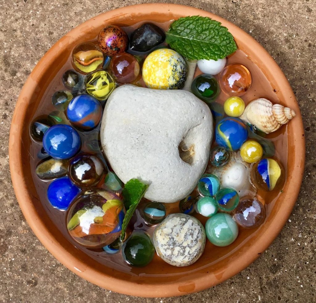 A terracotta dish filled with colorful marbles, a smooth stone, and a small seashell, surrounded by water and a mint leaf.