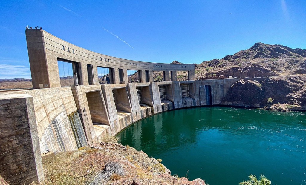 A concrete dam structure curved along a body of water, with visible gates and mountains in the background under a clear blue sky.