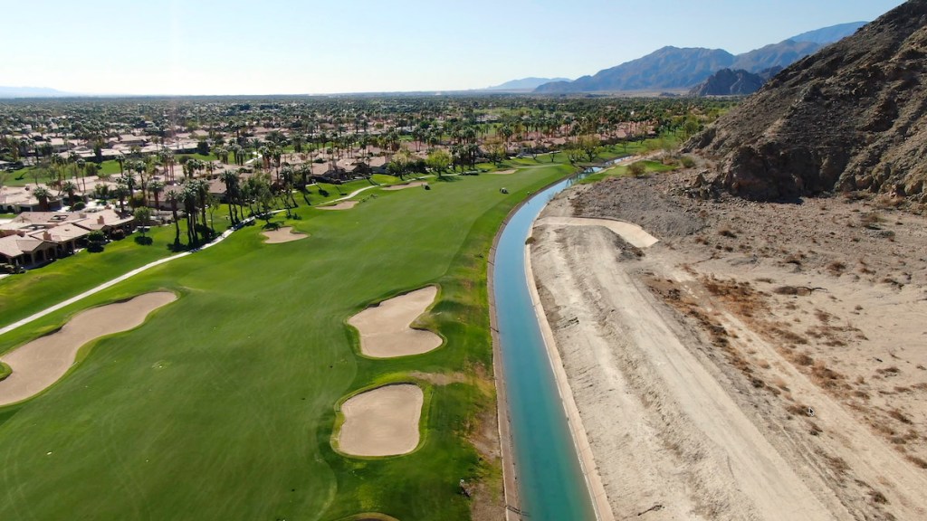 Aerial view of a golf course with well-maintained grass, sand traps, and a winding waterway, surrounded by residential homes and mountains in the background.