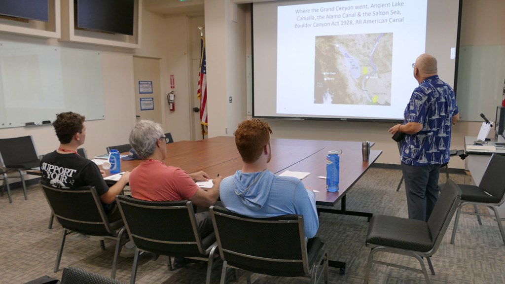 A classroom setting with four individuals seated at a large table, attentively listening to a presentation about the Grand Canyon, displayed on a screen. The presenter, wearing a patterned shirt, stands at the front of the room beside a map of the Grand Canyon's historical geography.
