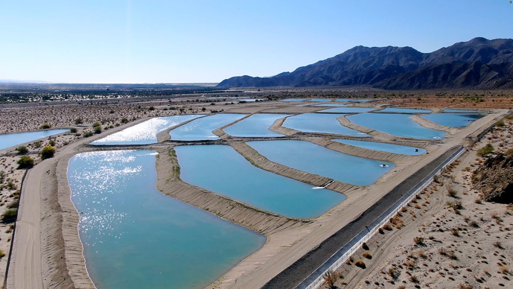 Aerial view of rectangular water ponds arranged in a grid pattern, surrounded by dry land and mountains in the background under a clear blue sky.