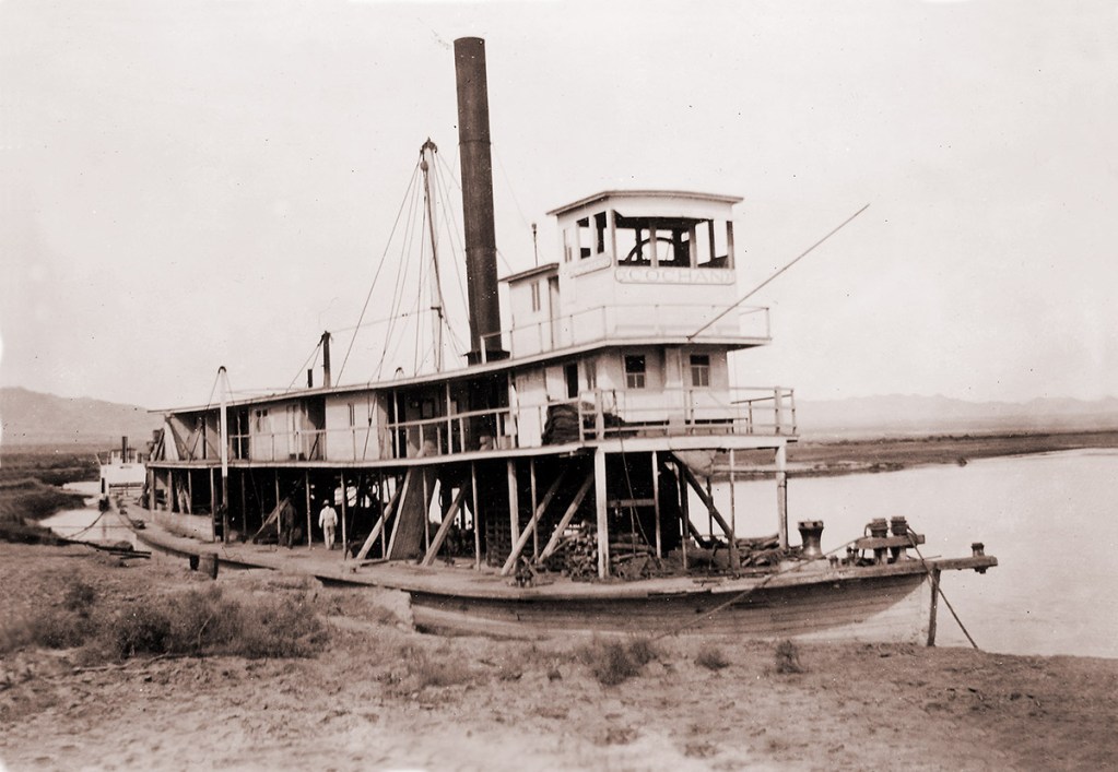 Historic steamboat docked near a riverbank, featuring a tall smokestack and a multi-level structure.