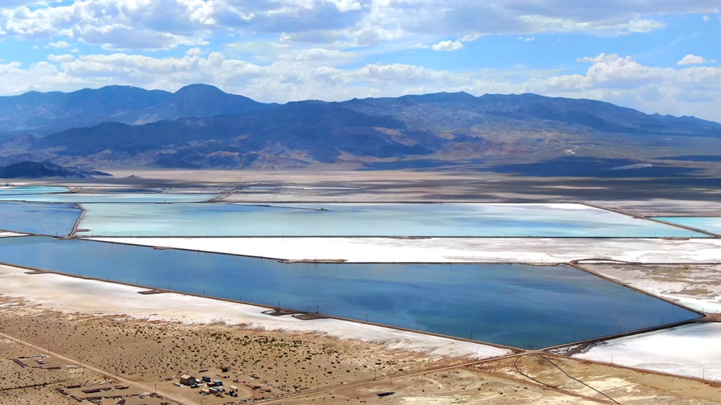 Aerial view of expansive salt flats with shallow ponds in various shades of blue and white, surrounded by desert terrain and distant mountains under a partly cloudy sky.