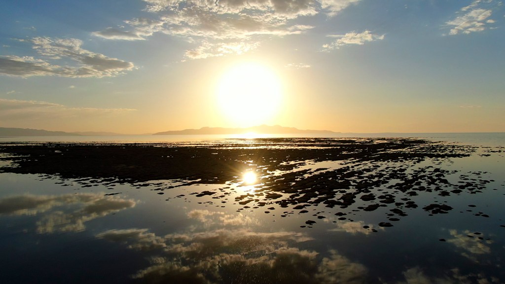 A serene sunset over a calm sea, with the sun reflecting on the water and rocky shoreline. The sky is filled with light clouds, and distant mountains are visible on the horizon.
