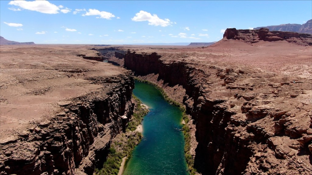 Aerial view of a winding river flowing through a deep canyon in a desert landscape under a blue sky with clouds.