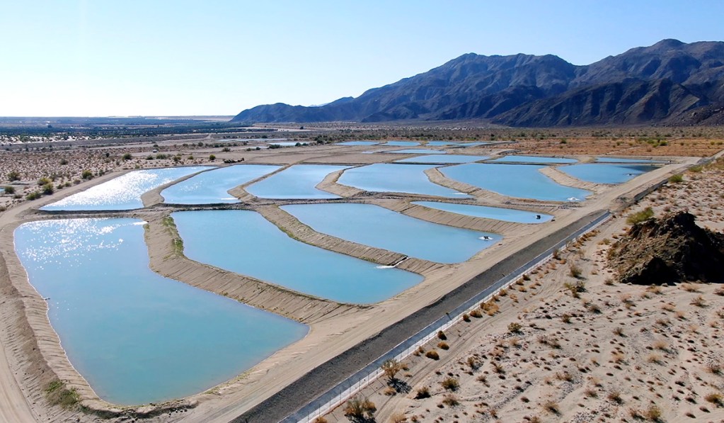 Aerial view of rectangular water ponds surrounded by sandy terrain and hills in the background, under a clear blue sky.