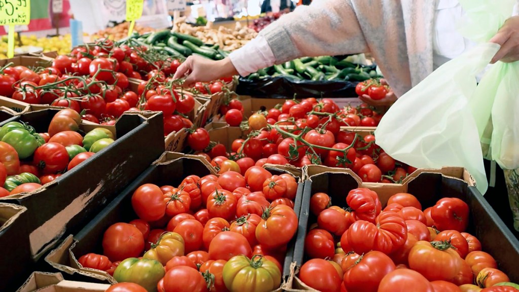 A customer selecting ripe tomatoes from a market stall, surrounded by various green and red produce in wooden crates.
