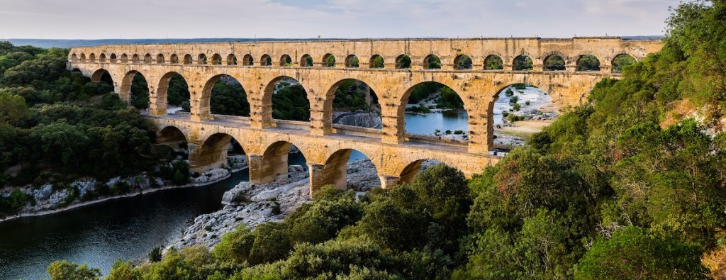 A view of the Pont du Gard aqueduct bridge spanning over a river, surrounded by lush greenery and rocky terrain.