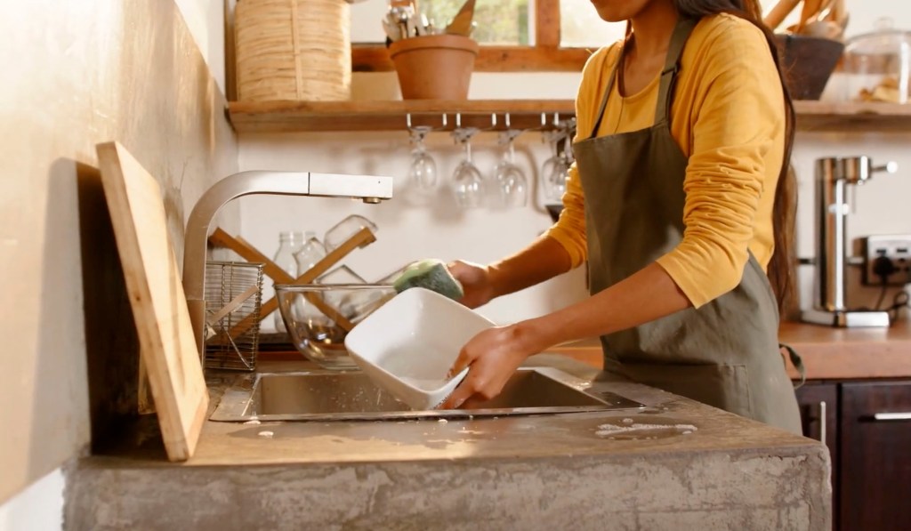 A person wearing an apron washes dishes in a modern kitchen sink, with various kitchen utensils and plants in the background.